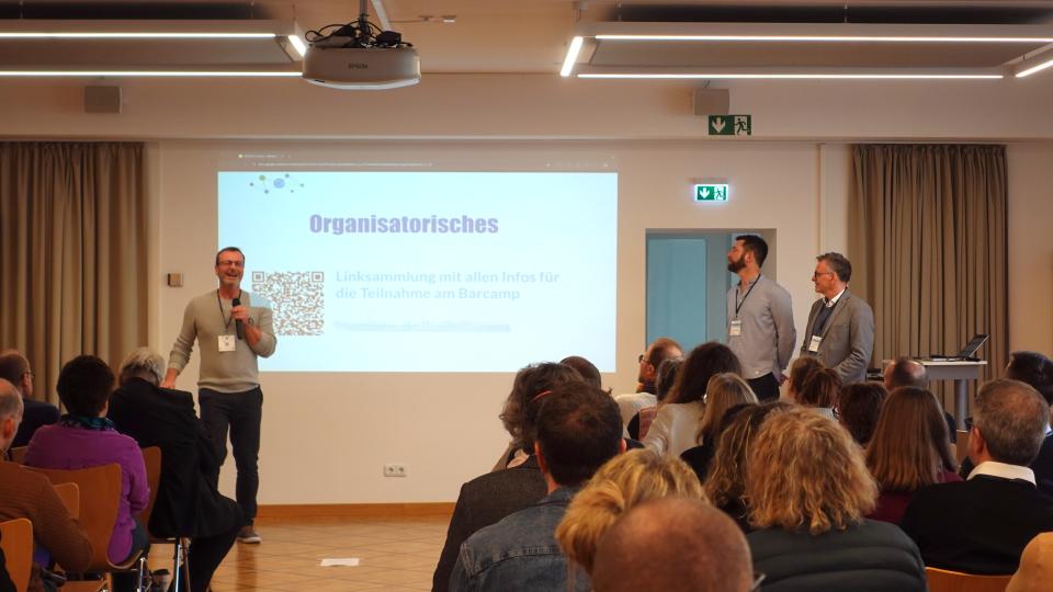 Die Verantwortlichen begrüßen die Teilnehmenden vor einer Power-Point-Präsentation.